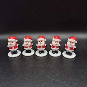 Könnte beinhalten: Fünf Miniatur-Weihnachtsmannfiguren. Jede Figur trägt einen roten Anzug mit weißer Verzierung, einen schwarzen Gürtel und einen passenden roten Hut. Sie stehen auf kleinen, weißen, kreisförmigen Sockeln, ideal für Weihnachtsdekorationen.