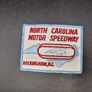 May include: A rectangular embroidered patch with a light blue border on a cream background. It features the red text NORTH CAROLINA MOTOR SPEEDWAY and ROCKINGHAM, N.C. A red oval track graphic surrounds a blue eagle silhouette on the patch.