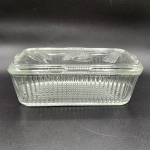 May include: A clear glass rectangular baking dish with a matching lid. The dish has vertical ribbed sides and a decorative design on the lid. The dish is sitting on a dark surface.