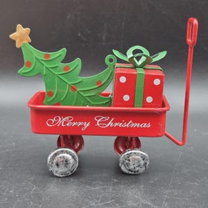 May include: A red metal wagon with the words "Merry Christmas" in white script. The wagon carries a green Christmas tree with red ornaments and a gold star, and a red gift box with a green bow. The wagon has four wheels and a red handle.