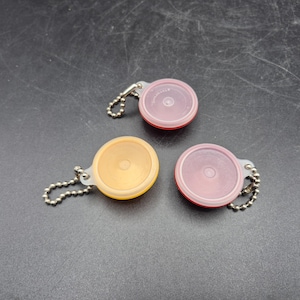 May include: Three circular, translucent plastic charms with metal ball chains attached. One charm is yellow, one is red, and one is a darker red or maroon. Each charm features a small, raised circular center and a clear, flexible plastic loop.