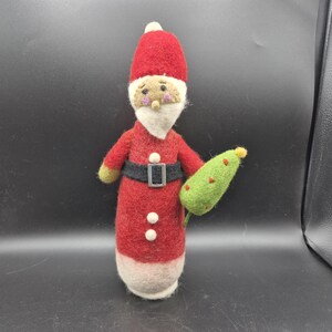 May include: A handmade felt Santa Claus figure, wearing a red suit with a black belt and white trim. The figure has a white beard, a red hat, and is holding a green Christmas tree. A festive holiday decoration.