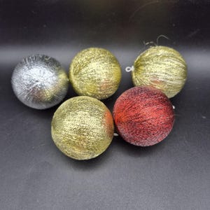 Op de afbeelding: Vijf decoratieve kerstballen. De bolvormige ornamenten zijn bedekt met glitter. Kleuren zijn zilver, goud en rood. Elk ornament heeft een kleine witte lus om op te hangen. Op een zwart oppervlak.