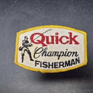 May include: A rectangular embroidered patch with a yellow border and white background. It features a black silhouette of a person fishing, the red text Quick, and the black text Champion FISHERMAN in a mix of script and block lettering.