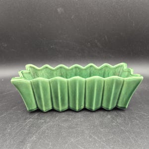 Puede incluir: Una jardinera rectangular de cerámica verde con un borde festoneado y lados verticales acanalados. La jardinera tiene un acabado brillante y una forma ligeramente acampanada, adecuada para plantas pequeñas o suculentas.