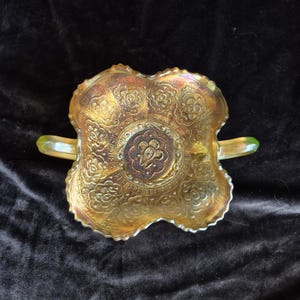 Puede incluir: Un cuenco iridiscente de estilo antiguo, con forma de cuatro lóbulos y dos asas. El cuenco presenta un patrón floral y geométrico en relieve en tonos dorados, bronce y verde. Las asas tienen puntas verdes.
