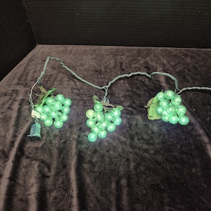 May include: A string of decorative lights featuring three clusters of green, textured, spherical bulbs shaped like grapes. Each cluster includes green fabric leaves attached to a dark green wire, with a standard electrical plug at one end.