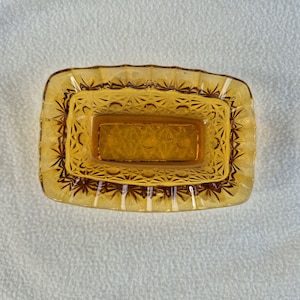 Op de afbeelding: Een rechthoekige, amberkleurige glazen schaal met een decoratieve, geschulpte rand. De schaal heeft een geometrisch patroon en een rechthoekig interieur. Het glas heeft een warme, gouden tint, wat op vintage duidt.