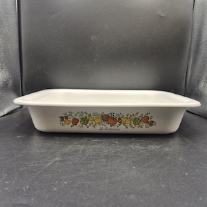 Puede incluir: Una fuente de horno rectangular de cerámica blanca con un borde ancho. El lado exterior presenta una colorida ilustración botánica de varias frutas y verduras, incluyendo tomates y alcachofas, con el texto Le Romarin impreso debajo.
