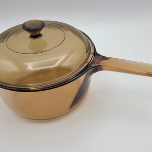 May include: Amber-colored glass saucepan with a matching lid. The saucepan has a long handle and a rounded body. The lid has a small knob for easy handling. The cookware is made of transparent glass.