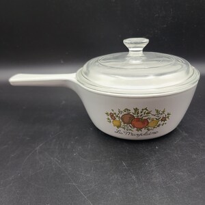 Puede incluir: Cazuela de cerámica blanca con tapa de vidrio y asa. La cazuela tiene un diseño floral con un tomate, un limón y hierbas. El texto "La Marjolaine" está impreso en la parte inferior de la cazuela.