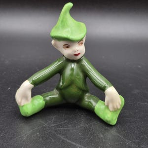 May include: A ceramic figurine of a seated elf with a green outfit and hat. The elf has a fair complexion, red lips, and is posed with legs spread and arms outstretched. The figurine is on a dark surface.