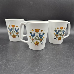 May include: Three white ceramic mugs with a decorative floral and bird design in orange, blue, and yellow. Each mug has a handle and a slightly flared shape, suitable for hot beverages. The mugs are arranged on a dark surface.