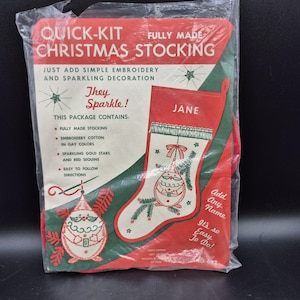 May include: A Quick-Kit fully made Christmas stocking craft package in a clear plastic bag. The red and white packaging features text, a graphic of a stocking with the name Jane, and illustrations of ornaments. It includes embroidery cotton, gold stars, and red sequins.