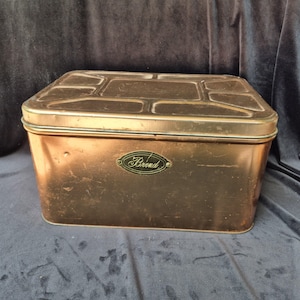 May include: A rectangular copper-colored metal storage tin with a lid featuring a geometric embossed pattern. The front of the container displays a small, dark oval plaque with the word Brend written in a cursive script, showing signs of wear.
