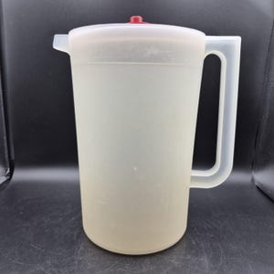 May include: A translucent, semi-clear plastic pitcher featuring a cylindrical body, a sturdy side handle, and a pouring spout. The pitcher is topped with a matching translucent lid that includes a small, circular, solid red knob at the center.