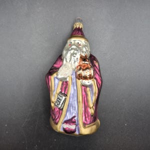 May include: A glass Christmas ornament shaped like a figure with a long white beard, wearing a deep magenta robe with gold trim and a light purple inner garment. The figure holds a small brown owl and a white textured object, with a metal hanging loop.