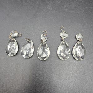 Pode incluir: Cinco pingentes de lustre de cristal transparente com formatos de gota facetados e pequenas peças superiores redondas. Cada pingente tem um gancho de metal para pendurar. Os pingentes estão dispostos sobre uma superfície cinza escuro.