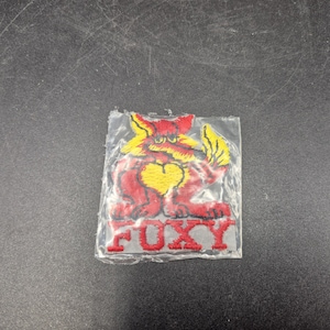 May include: A rectangular embroidered patch encased in clear plastic featuring a red and yellow fox character with a heart on its chest. The word FOXY is stitched in red capital letters at the bottom of the design, which is set against a light gray fabric background.