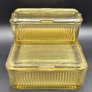Puede incluir: Dos recipientes rectangulares vintage de vidrio amarillo para refrigerador con lados acanalados y tapas a juego. Los recipientes están apilados, mostrando su diseño y color. Las tapas tienen una pequeña pestaña para facilitar la apertura.