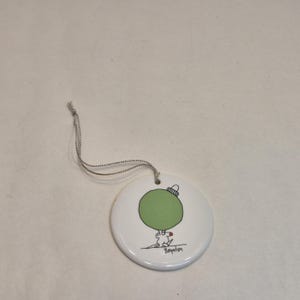 Op de afbeelding: Een witte keramische kerstversiering met een groen ornamentontwerp en een zilveren draad om op te hangen. De versiering heeft een cartoonillustratie van een figuur die het groene ornament vasthoudt. Het woord "Boynton" staat eronder.