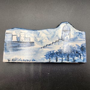May include: A rectangular ceramic piece with a hand-painted landscape scene in shades of blue and white. The design includes buildings, a domed structure, trees, and the text "F St. Anna".