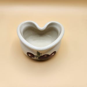 Könnte beinhalten: Eine weiße, herzförmige Schale aus Keramik mit einem braunen und grünen Apfeldesign. Die Schale ist leer und bereit zum Befüllen.