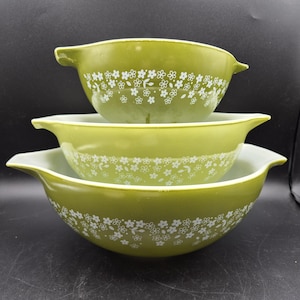 Op de afbeelding: Een set van drie vintage groene Pyrex mengkommen. Elke kom heeft een wit bloemenpatroon aan de buitenkant. De kommen zijn genest, met de grootste kom onderaan en de kleinste bovenaan. De kommen hebben handvatten.