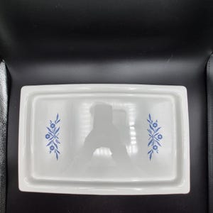 Puede incluir: Un plato rectangular de cerámica blanca con borde elevado. El plato presenta un estampado floral azul en cada extremo. El diseño incluye flores y hojas estilizadas. El plato es probablemente para servir o exhibir.