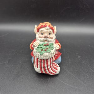May include: A ceramic Christmas elf figurine. The elf is wearing a red and yellow hat and coat, with a white beard and holding a stocking filled with holly. The stocking is white with red stripes.