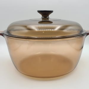Puede incluir: Una fuente para horno de vidrio transparente de color ámbar con tapa a juego. La fuente tiene dos asas laterales y un pomo marrón oscuro en la tapa. El utensilio de cocina está diseñado para cocinar y servir.