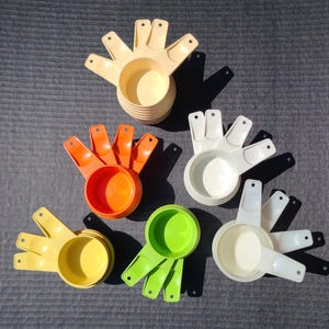 May include: Set of six nesting measuring cups in various colours, including white, orange, yellow, and green. The cups are made of plastic and have a hand-shaped design.
