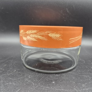 Op de afbeelding: Een heldere glazen pot met een ronde basis en een brede, oranje deksel. De deksel is versierd met een tarwepatroon in goud. De pot is waarschijnlijk voor het bewaren van voedsel.