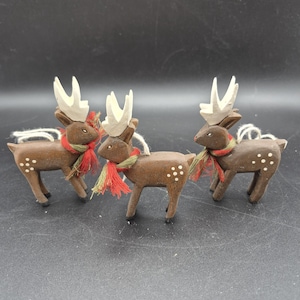 Puede incluir: Tres adornos de renos de madera. Cada reno tiene astas blancas, una bufanda roja y verde y detalles de puntos blancos. Están ensartados con una cuerda blanca para colgar. Los renos son una decoración festiva navideña.
