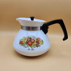 May include: White enamel coffee pot with a black handle and lid. The pot has a floral design with fruit and the text "Le Thé" on the front.