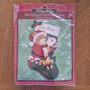 Könnte beinhalten: Ein Filz-Applikations-Weihnachtsstrumpf-Set in einer transparenten Kunststoffverpackung. Das Design zeigt einen Teddybären als Weihnachtsmann, der eine Santa-Figur hält. Der Strumpf ist grün mit roten und weißen Akzenten. Die diagonale Größe beträgt 45,7 cm.