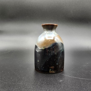 May include: A small, handcrafted ceramic vase with a unique glaze. The vase features a dark brown rim, transitioning to a light brown and blue-grey body, and a dark, speckled base. The bottle-shaped vase is suitable for displaying small flowers or as a decorative piece.