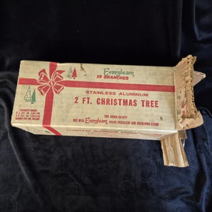 May include: A rectangular cardboard box for an Evergleam 2 ft. stainless aluminum Christmas tree with 19 branches. The box features red ribbon graphics, green tree illustrations, and text detailing patent numbers and usage instructions.