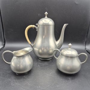 May include: A vintage pewter tea set, including a teapot with a woven handle, a creamer, and a lidded sugar bowl. The set has a classic design with a matte finish. The teapot is approximately 9 inches tall.