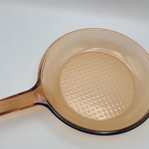 Puede incluir: Sartén de vidrio color ámbar con un mango largo. El interior de la sartén tiene un patrón de diamantes. La sartén está diseñada para cocinar en la estufa y está hecha de vidrio duradero.