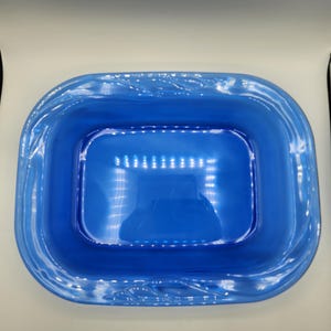 Könnte beinhalten: Eine leuchtend blaue, rechteckige Glasschale mit abgerundeten Ecken. Die Schale hat einen leicht erhöhten, strukturierten Rand. Das Innere ist glatt und reflektiert das Licht, wodurch ein glänzendes Aussehen entsteht. Das Design ist schlicht und elegant.