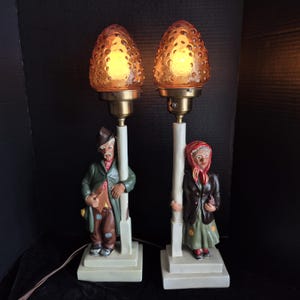 May include: Pair of vintage table lamps featuring figures of a man and woman leaning on posts. The lamps have textured amber-colored glass shades and brass fittings. The figures are painted in various colors, including green, brown, and red.