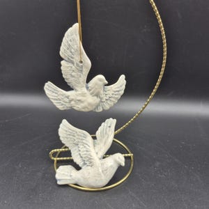 May include: Two white dove ornaments with outstretched wings, suspended by gold-colored strings. The doves are perched on a gold-colored metal ring. The background is a dark gray color.