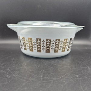 May include: A white casserole dish with a clear glass lid. The dish features a gold leaf pattern around the middle. The handles are white, and the dish is sitting on a dark surface. A vintage kitchenware item.