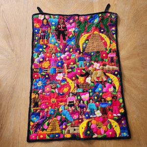 May include: A vibrant, embroidered textile featuring a complex design with figures, flowers, and geometric shapes in a variety of colors. The artwork is framed with a black border and includes hanging loops.