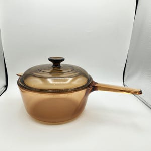 May include: Amber-colored glass saucepan with a matching lid and a long handle. The saucepan has a rounded shape and a clear glass lid with a small knob. The handle is also made of glass. This cookware is suitable for stovetop use.
