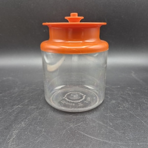 Op de afbeelding: Een doorzichtige plastic pot met een oranje deksel en een klein handvat aan de bovenkant. De pot heeft een cilindrische vorm en is leeg. Het deksel past goed op de pot.