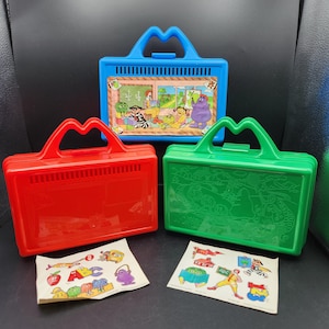 Pode incluir: Três lancheiras de plástico com personagens de desenhos animados coloridos e letras. As lancheiras são vermelha, azul e verde. Cada lancheira tem uma alça e um fecho.