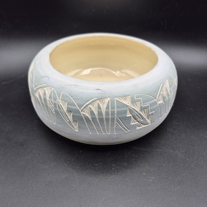 Puede incluir: Un cuenco redondo de cerámica azul claro con interior crema. El exterior presenta un diseño geométrico blanco tallado. El cuenco está sobre una superficie negra.