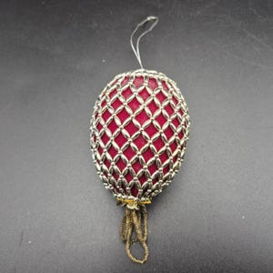 May include: A decorative egg-shaped ornament with a red velvet interior and a silver-colored, beaded overlay. The ornament has a small loop for hanging and a beaded tassel at the bottom. The ornament is approximately 3 inches tall.